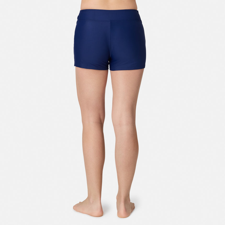 A person stands barefoot on a plain white background, facing away from the camera and wearing the Free Country Women's Fully Lined Swim Skort in navy blue, offering comfort and coverage.
