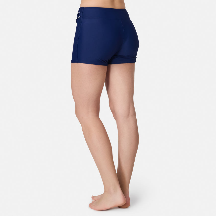 A woman stands barefoot on a white background, shown from the back, wearing the Free Country Women's Fully Lined Swim Skort in navy blue.