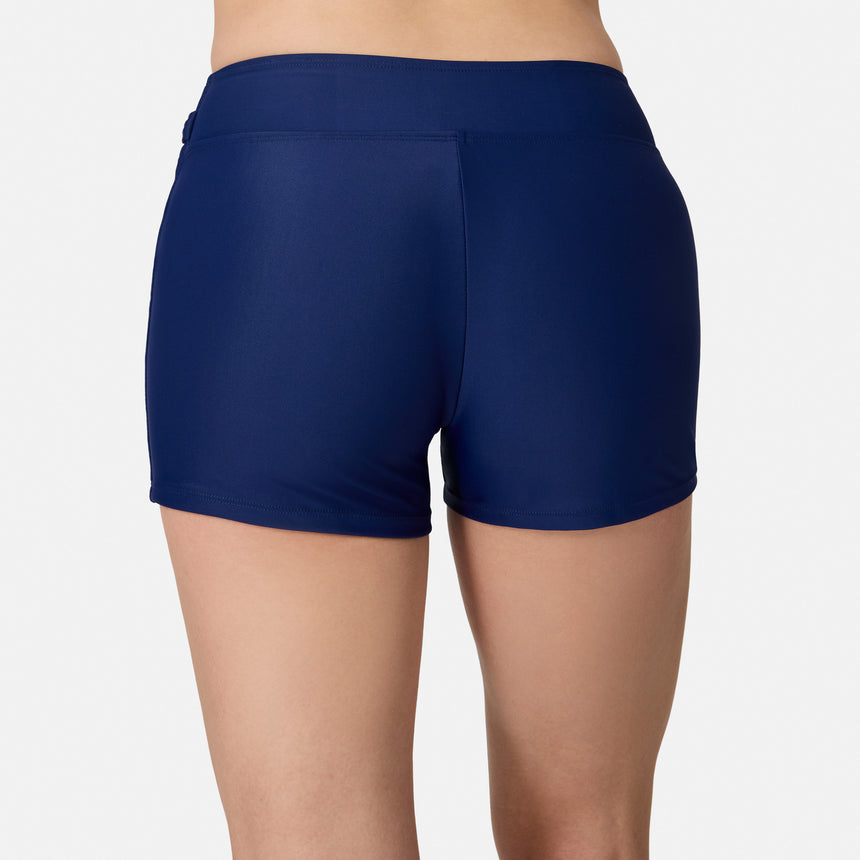 A person is shown from the waist down, facing away from the camera, modeling the Free Country Women's Fully Lined Swim Skort in navy blue. This swim bottom provides UPF 50+ protection and is set against a plain white background.