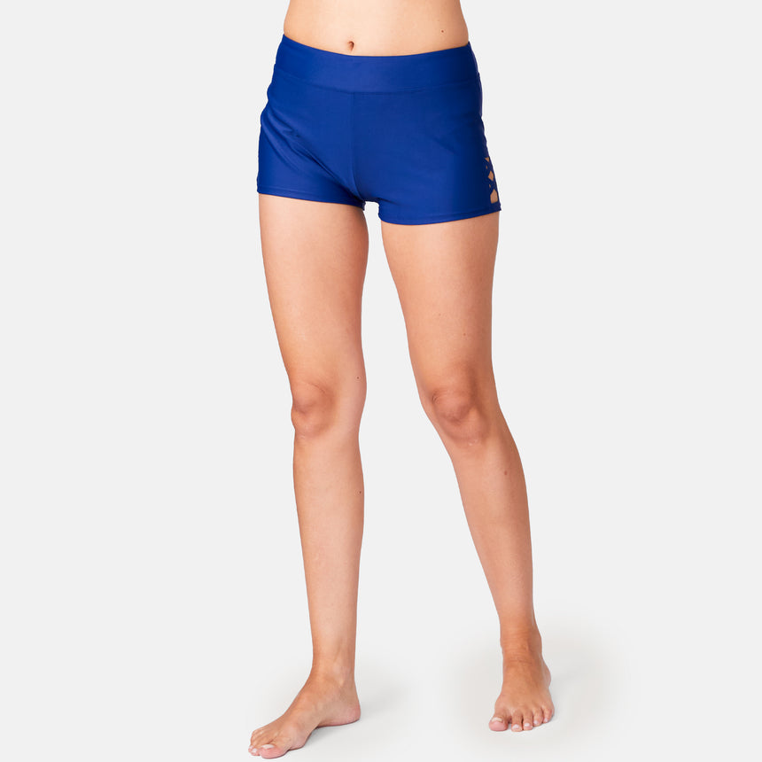 A person stands barefoot against a plain light background, modeling Free Country Women's Laser Cut Boyshorts in royal blue, featuring a snug fit, high waistband, and sustainable recycled materials. Only the legs and lower torso are visible.