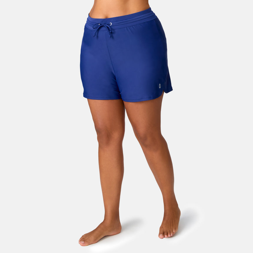A woman stands barefoot in Free Country Women's Plus Size Drawstring Swim Short, shown from mid-torso to feet against a plain light background.