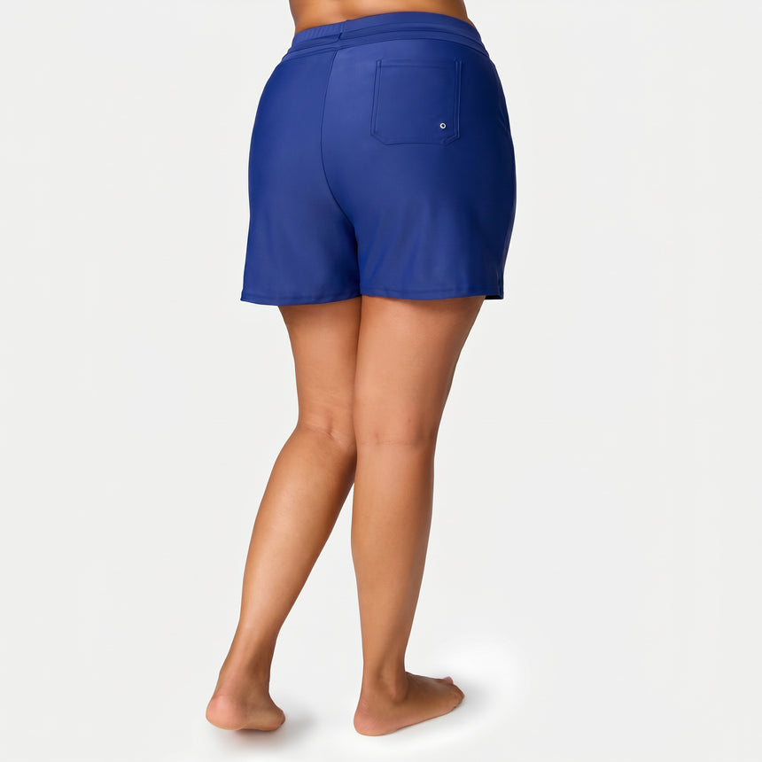 A woman stands barefoot, facing away, wearing Free Country Women's Plus Size Drawstring Swim Short in royal blue with a single back pocket and UPF 50+ protection, set against a plain light gray background.