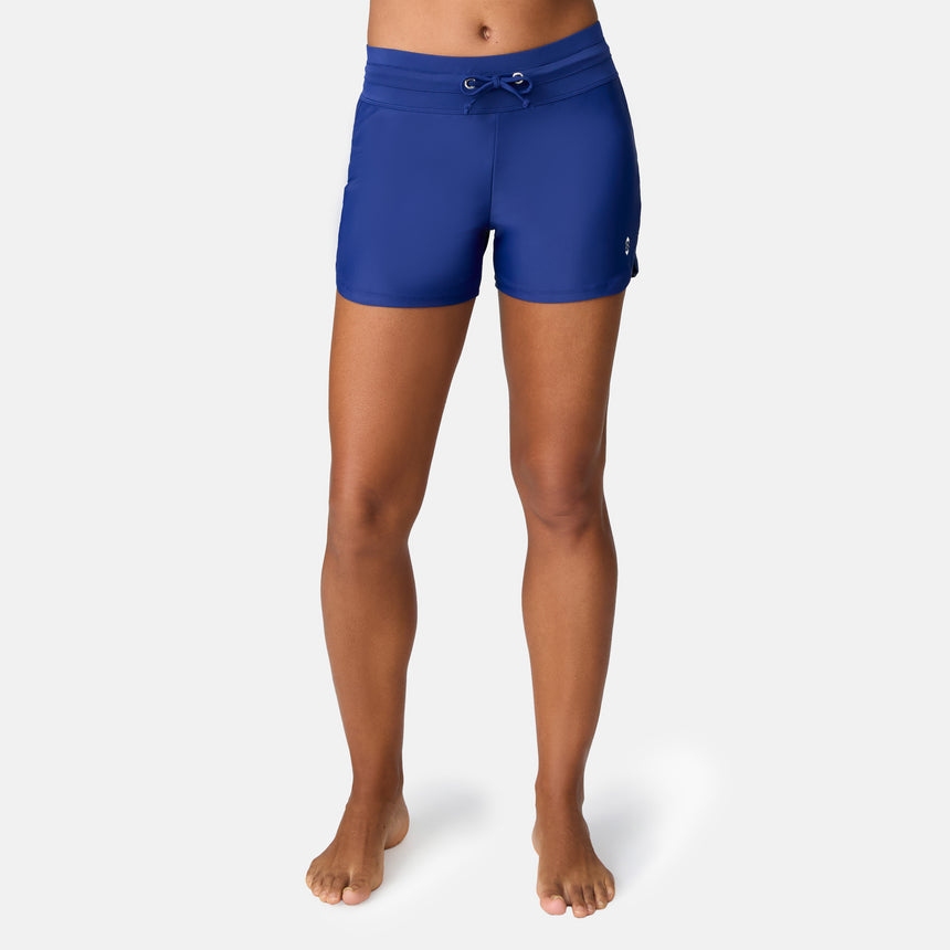 A person stands barefoot against a plain backdrop, modeling Free Country Women's Drawstring Swim Shorts in navy with a mid-thigh cut and UPF 50+ protection. Their torso is not shown.