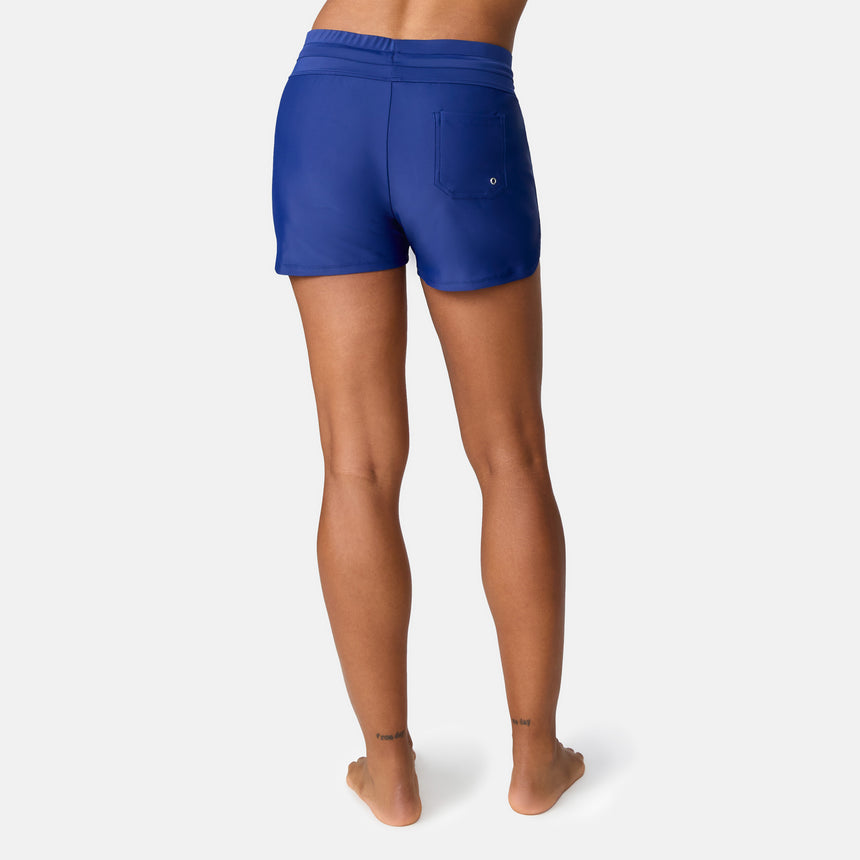 A woman stands barefoot, facing away, wearing Free Country Women's Drawstring Swim Short in royal blue with a back right pocket. These quick-dry shorts offer UPF 50+ protection, set against a plain white background.