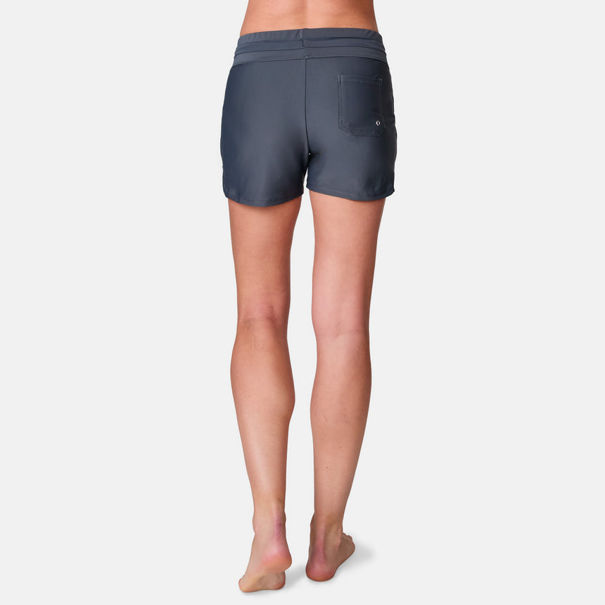 A woman stands barefoot facing away, wearing Free Country Women's Drawstring Swim Short in dark grey with a back pocket and UPF 50+ protection, shown against a plain white background.