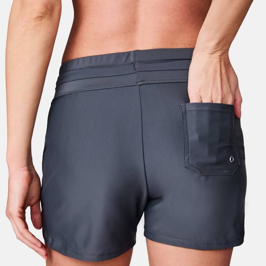 A woman wearing Free Country Women's Drawstring Swim Shorts in dark gray stands with her back to the camera, one hand partially tucked into the back pocket of the quick-dry shorts.