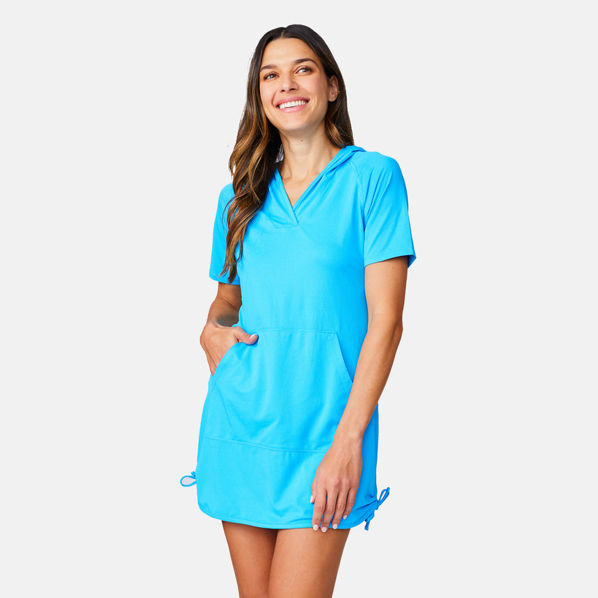 A woman smiles in the Free Country Women's SunFree UPF Dress, a bright blue short-sleeve hooded dress with UPF 50+ protection, front pockets, and hem drawstrings, standing against a plain light background.