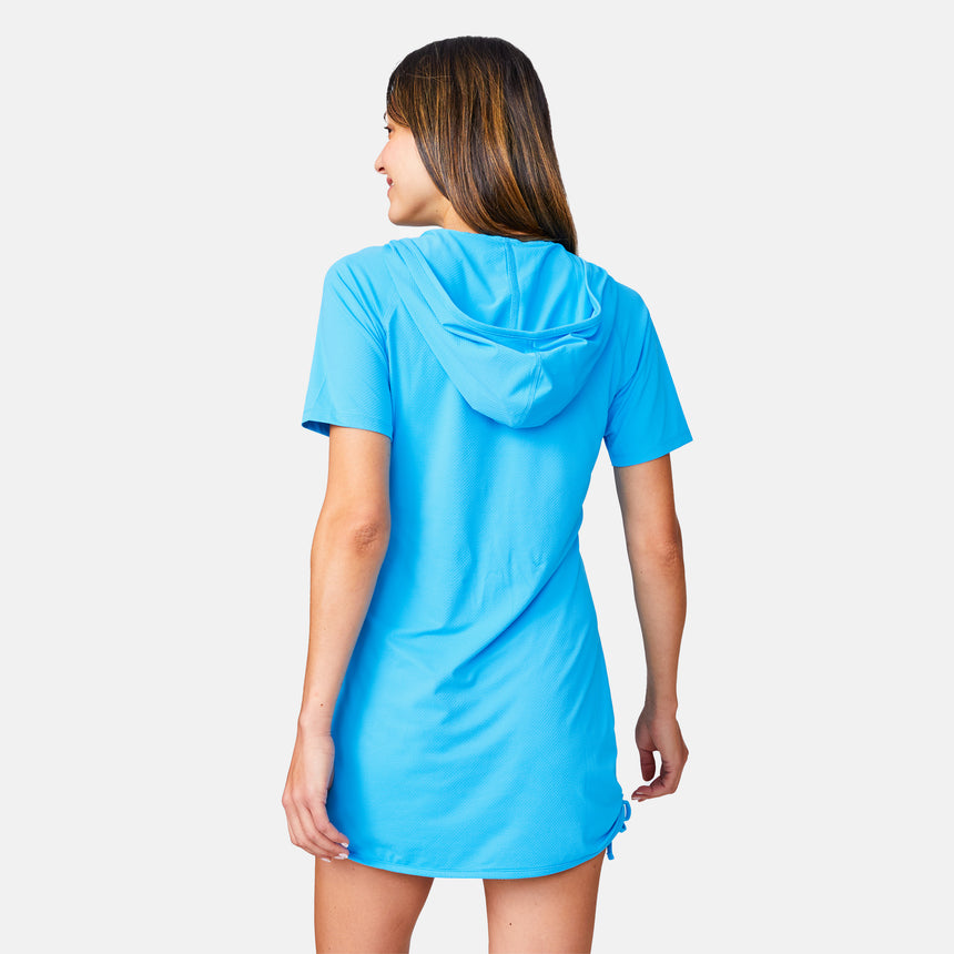 A woman with long brown hair faces away, wearing the Free Country Women's SunFree UPF Dress— a bright blue, short-sleeve hooded cover-up made from recycled polyester. The background is plain white.