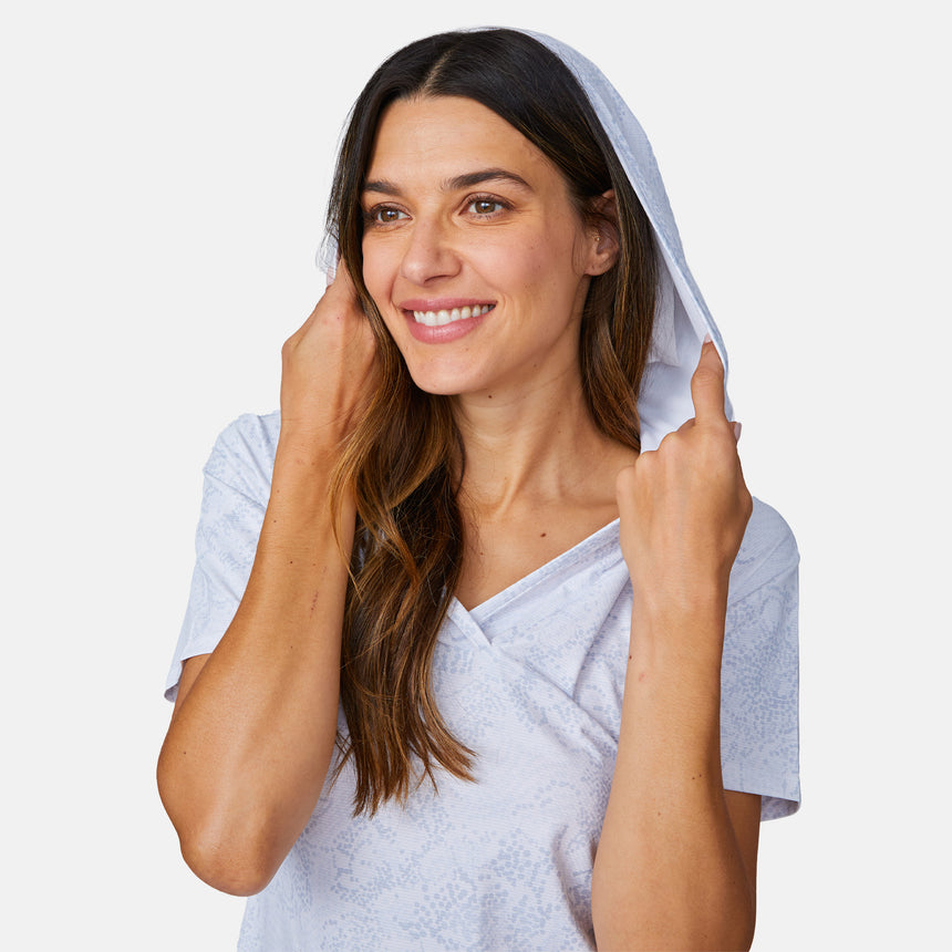 A smiling woman with long brown hair wears the Free Country Women's SunFree UPF Dress, a light-colored, short-sleeve hoodie cover-up made from recycled polyester, holding the hood with both hands against a plain white background.