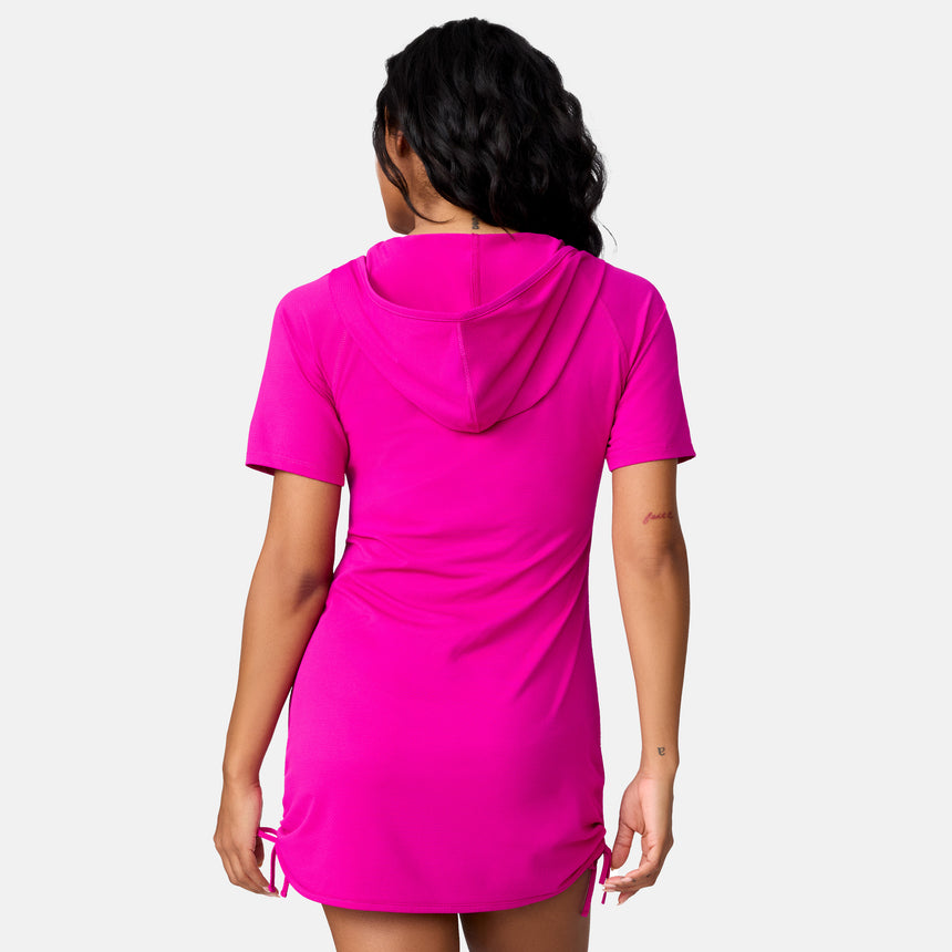 A woman with loose wavy dark hair models the Free Country Women's SunFree UPF Dress—a bright pink, short-sleeve, hooded dress with side drawstrings—while standing against a plain white background.