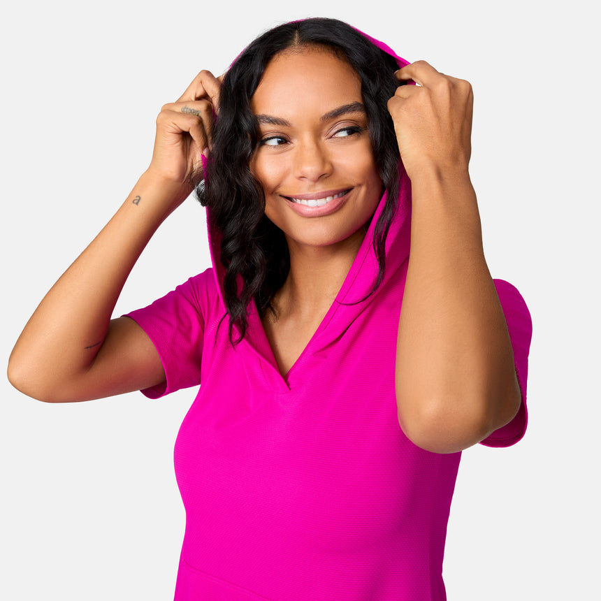 A woman with wavy hair smiles while holding the hood of a bright pink Free Country Women's SunFree UPF Dress, made from recycled polyester, against a plain light background.