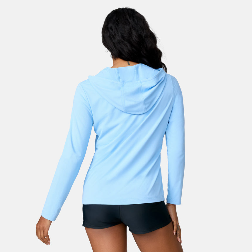 A woman faces away from the camera, wearing the Free Country Women's SunFree Full Zip UPF Sunshirt in light blue with black shorts, posed against a plain white background.