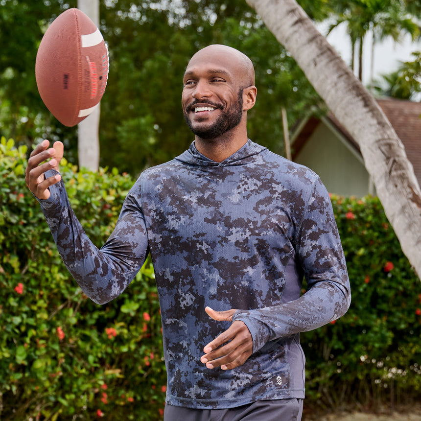 Man wearing Free Country Men's SunFree UPF Printed Hoodie in Charcoal Camo, size M at the park, made from anti-odor, breathable lightweight recycled polyester performance fabric featuring a classic active fit, styled with grey shorts #color_charcoal-camo