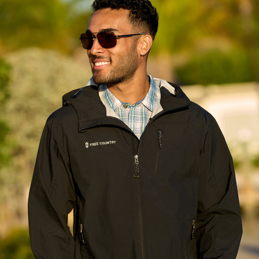 Man wearing Free Country Men's Hydro Lite X2O Status Jacket in Black, size M, made from lightweight four way stretch water-resistant performance fabric featuring tab cuffs, an attached hood with a drawcord, and a chest and lower pockets, styled with a plaid shirt #color_black