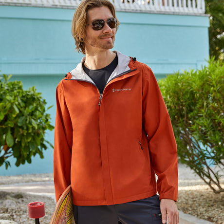 Man wearing Free Country Men's Hydro Lite X2O Spectator Waterproof Jacket in Harris Orange, size M, made from lightweight four way stretch performance fabric featuring sealed seams, elastic cuffs, and an attached hood with a drawcord, styled with blue jeans #color_harris orange