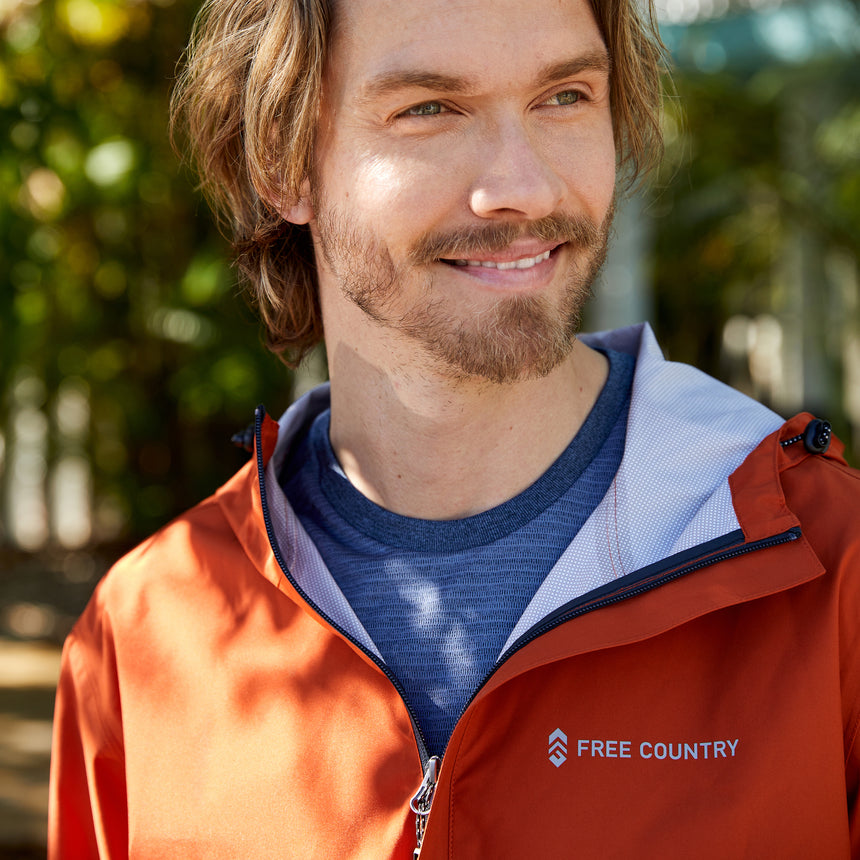 Man wearing Free Country Men's Hydro Lite X2O Spectator Waterproof Jacket in Harris Orange, size M, made from lightweight four way stretch performance fabric featuring sealed seams, elastic cuffs, and an attached hood with a drawcord, styled with a blue t shirt #color_harris orange