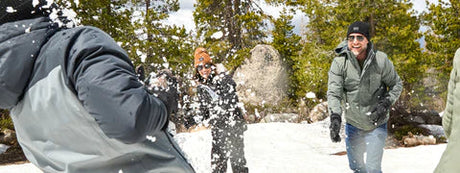 Friends in winter jackets and beanies walk through a snowy landscape with trees and rocks.