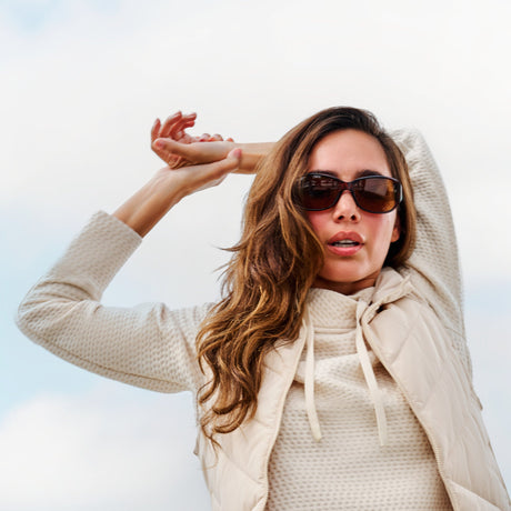 Woman wearing sunglasses and a light-colored jacket poses with her arms raised above her head against a bright, cloudy sky background.