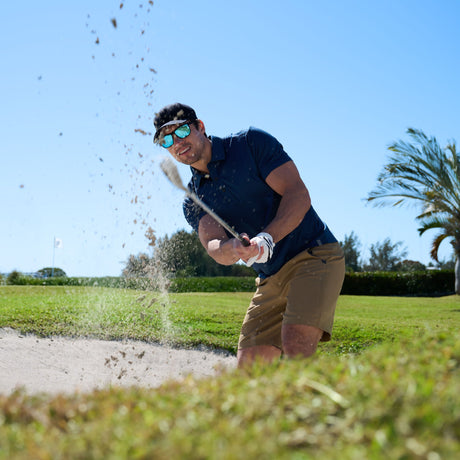 A man wearing sunglasses and golf gloves hits a golf ball out of a sand bunker on a sunny day, with sand flying into the air and a palm tree in the background.