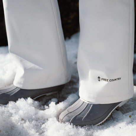 Close-up of a person standing on snow wearing gray waterproof boots and white pants with the logo FREE COUNTRY near the hem.