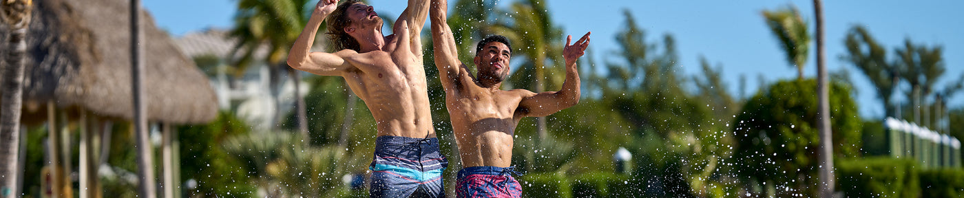 Two men in colorful Free Country men's swim board shorts run on the beach.