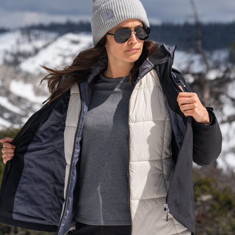 A woman wearing sunglasses, a gray beanie, and layered winter jackets stands outdoors in a snowy mountain setting, looking to the side with her jacket slightly open.