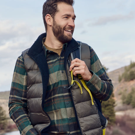 A smiling man with a beard wearing a green plaid shirt and a puffer vest stands outdoors holding a backpack strap, with trees and hills in the background.