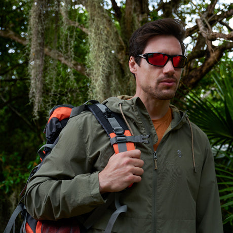 A man wearing red sunglasses and an olive green jacket carries a hiking backpack in a lush, green forest with moss hanging from tree branches in the background.