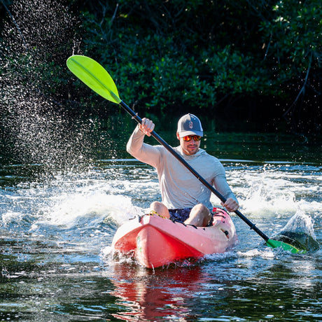 A man wearing sunglasses and a cap kayaks energetically on a river, causing water to splash up around him, with green foliage visible in the background.