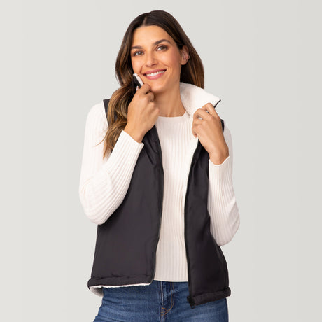 A woman with long brown hair smiles while holding the collar of the Women's Cascade Canvas 3-in-1 Systems Jacket by Clearance, featuring a white ribbed sweater and reversible inner vest, against a plain light gray background.