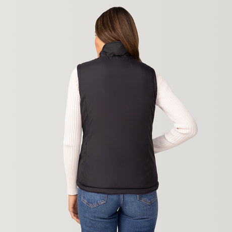 A woman with long brown hair, wearing the Clearance Women's Cascade Canvas 3-in-1 Systems Jacket over a white ribbed sweater and blue jeans, stands facing away from the camera against a plain light background.