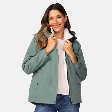A woman with long brown hair smiles while wearing the Clearance Women's Cascade Canvas 3-in-1 Systems Jacket in sage green over a white top and blue jeans, set against a light gray background. She holds the jacket's collar with both hands.