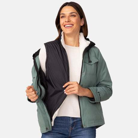 A woman with long brown hair smiles as she unzips the Women's Cascade Canvas 3-in-1 Systems Jacket by Clearance, wearing it over a white sweater against a plain light background.