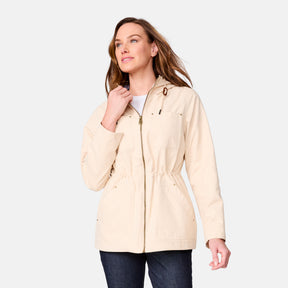 A woman wears the Clearance Women's Lightweight Cascade Canvas Jacket in light beige, featuring a front zipper and drawstring waist, over a white top with dark jeans, standing against a plain light background.