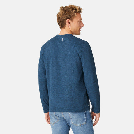 A man with short, curly blond hair wears a blue Men's FreeCycle® Sueded Spacedye Long Sleeve Crew Neck by Free Country and blue jeans. He faces away from the camera, glancing over his right shoulder against a plain light background.