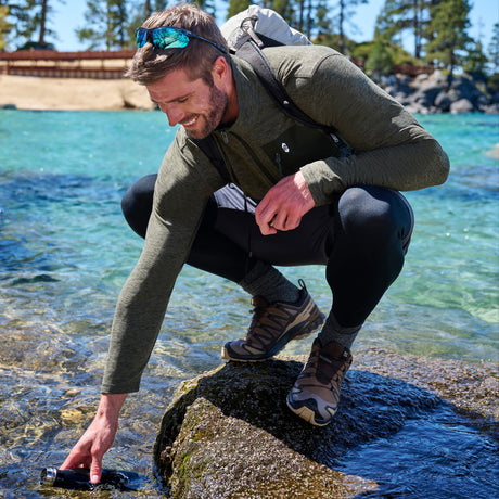 A man crouches on a rock by the ocean, reaching into the water for a bottle. He is wearing an olive green long-sleeve technical top, black leggings, hiking boots, and a backpack in a sunny outdoor coastal setting.