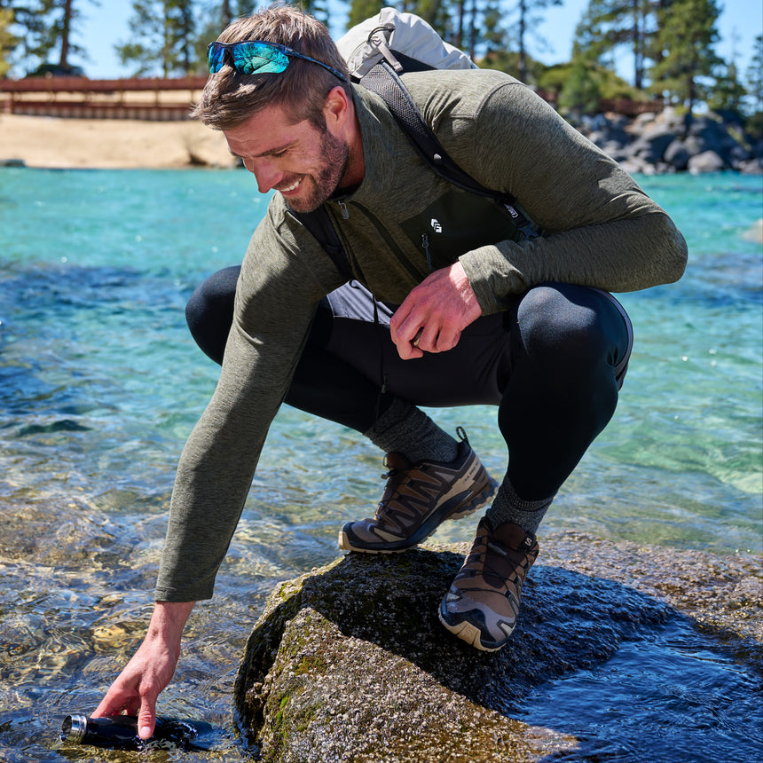 A man crouches on a rock by the ocean, reaching into the water for a bottle. He is wearing an olive green long-sleeve technical top, black leggings, hiking boots, and a backpack in a sunny outdoor coastal setting.