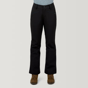 A person is shown from the waist down wearing Free Country Women's Stormlite Super Softshell® Ski Pant in black, paired with a gray shirt and brown lace-up boots, standing against a plain light background.