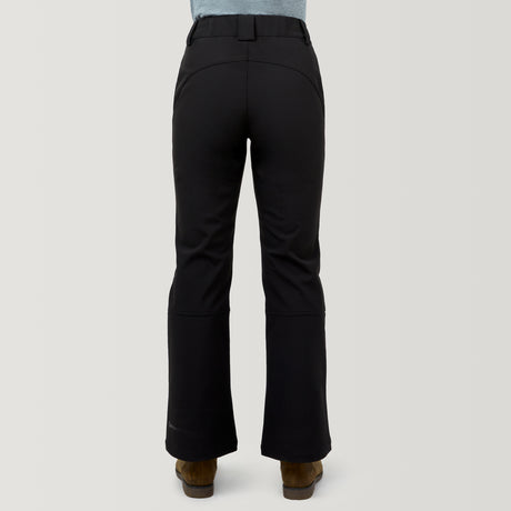 A person is shown from the back wearing Free Country Women's Stormlite Super Softshell® Ski Pants in black, paired with brown shoes, standing against a plain light gray background.