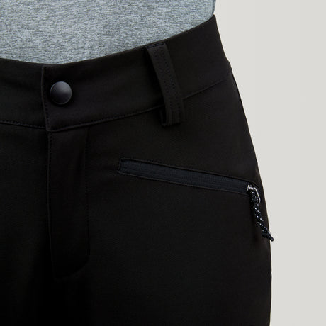 Close-up of a person wearing the Free Country Women's Stormlite Super Softshell® Ski Pant in black, featuring a zipper pocket and black waist button, paired with a tucked-in light gray shirt.