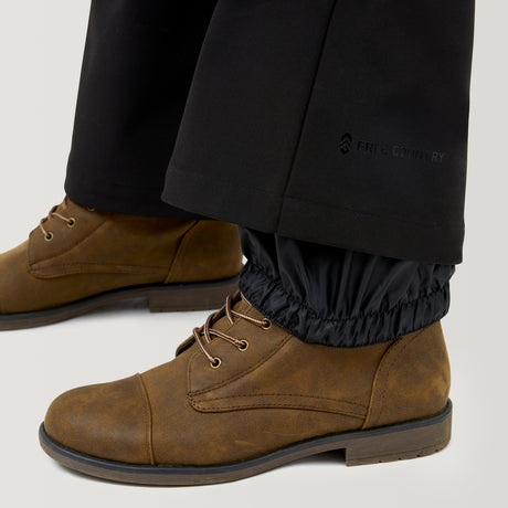 Close-up of a person wearing brown lace-up boots and black Women's Stormlite Super Softshell® Ski Pants by Free Country, featuring water resistance, elastic gaiters tucked into the boots, and Free Country branding on the lower leg.