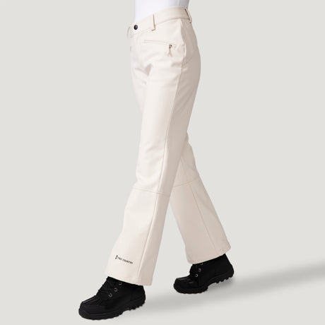 A person wears Free Country Women's Stormlite Super Softshell® Ski Pant in off-white with straight-leg fit, zippered pockets, four-way stretch, and a small ankle logo. Black outdoor shoes and a plain light gray background complete the outfit.