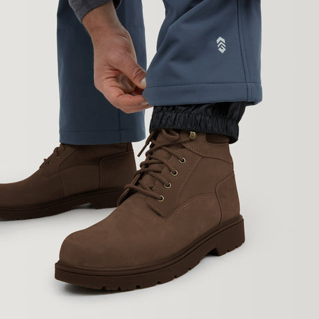 A person in Clearance Men's Ascend Flex Softshell Ski Pant and brown lace-up boots adjusts the elastic cuff of their black inner pants, tucked into the boots. The plain, light-colored background keeps focus on the outfit.