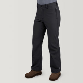 Wearing Men's Ascend Flex Softshell Ski Pant by Clearance in dark gray with zippered side pockets and tailored fit, paired with brown hiking boots. Water-resistant ski pants ideal for winter adventures. Person's upper body not visible, standing against a plain light background.