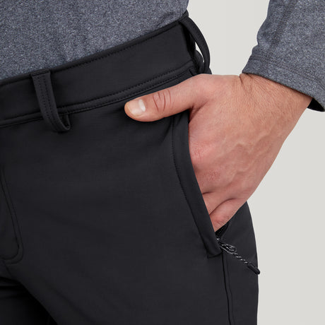 A person in a gray long-sleeve shirt places their right hand into the front pocket of black Clearance Men's Ascend Flex Softshell Ski Pant, showing the visible zipper pull.