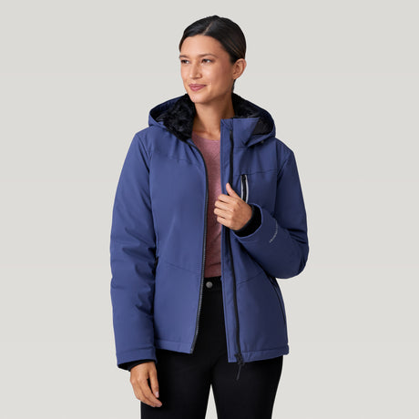 A woman models the Clearance Women's Thermo Super Softshell® Ski Jacket in blue over a mauve shirt, standing against a plain light gray background and smiling slightly as she looks off to the side.