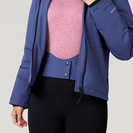A person wears the Women's Thermo Super Softshell® Ski Jacket by Clearance, partially unzipped to show a blue waist panel with snap buttons over a pink top and black pants. The water-resistant blue jacket is featured; the person’s head is not visible.
