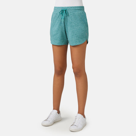A person is wearing Free Country Women's Cloud Knit Shorts in teal, featuring four-way stretch, a drawstring, moisture-wicking fabric, and curved hems. They pair the shorts with a white top and white sneakers against a plain white background.