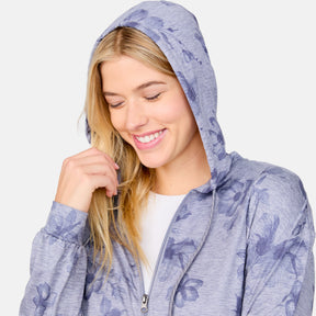 A woman with long blonde hair smiles while looking down, wearing the Clearance Women's Cloud Knit Zip Hoodie in light purple with a floral pattern over a white shirt. Her hood is up and she lightly touches her hair.