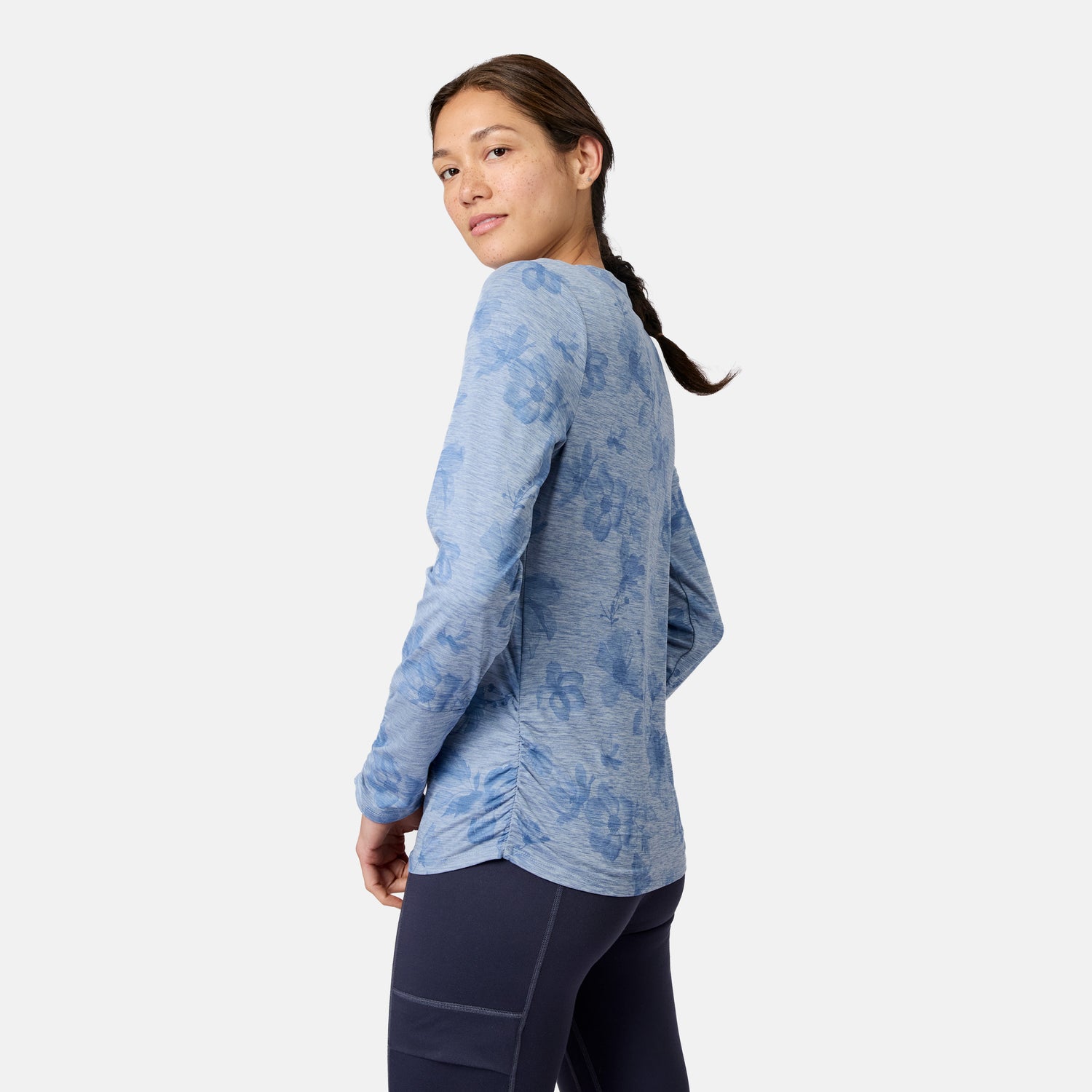 A woman in a Women’s Cloud Knit Crew Neck Top by Free Country and dark leggings stands sideways, looking over her shoulder against a plain white background. The blue-patterned shirt features moisture-wicking fabric.